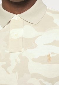 Beige polo tričko s jemným maskáčovým vzorem, texturovaná látka, tři knoflíky na límečku a vyšívané logo na hrudi.