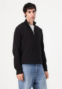 Pull à col zippé noir avec des poignets côtelés, fabriqué en tissu doux, associé à un jean en denim bleu clair. Le design présente un petit détail de logo.