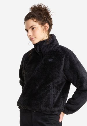 Frau mit lockigem Haar in einem hohen Dutt, trägt eine schwarze, flauschige Fleecepullover mit Viertel-Reißverschluss und schwarze Hose, schaut nach links, Hände in die Hüften gestützt.