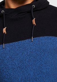 Sweat à capuche avec un haut texturé noir et une partie inférieure en tricot bleu, avec des accents de matériel beige et des cordons noirs.