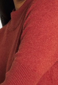 Detailní záběr na osobu v červeném pleteném svetru s žebrovanými manžetami a vysokým límcem, který ukazuje část krku a ramene.