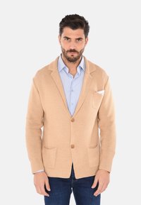 Cardigan in maglia beige con finitura testurizzata, due tasche anteriori e bottoni di legno. Indossato sopra una camicia azzurro chiaro, con un fazzoletto bianco nella tasca.