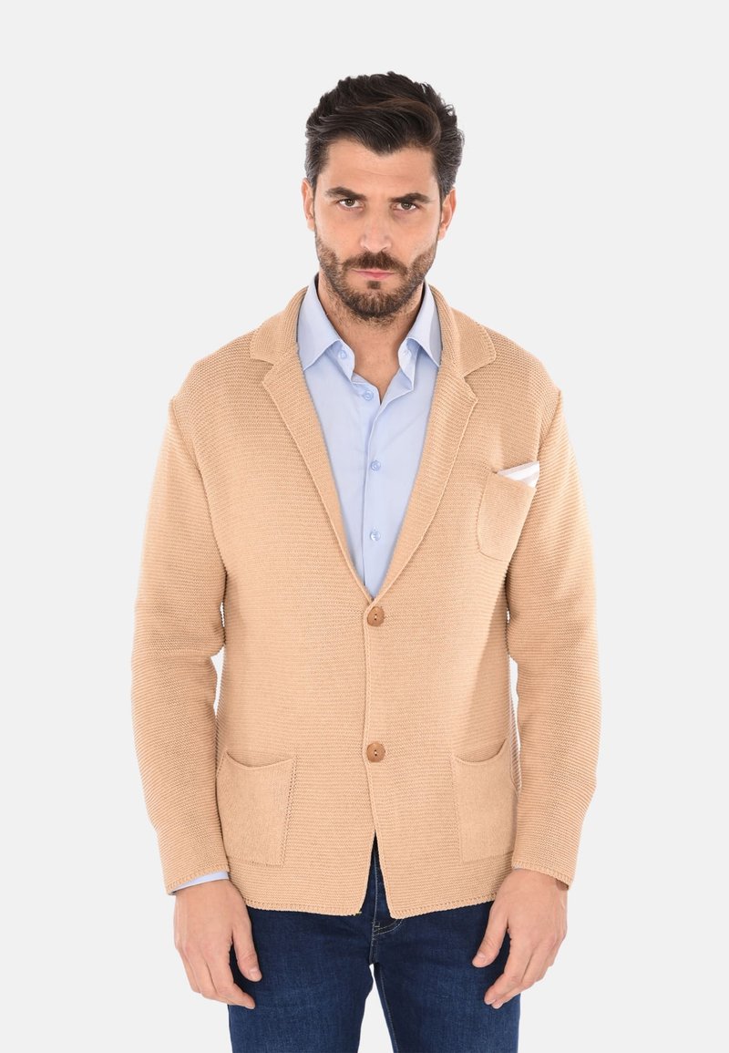Cardigan in maglia beige con finitura testurizzata, due tasche anteriori e bottoni di legno. Indossato sopra una camicia azzurro chiaro, con un fazzoletto bianco nella tasca.