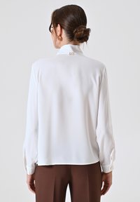 Witte blouse met lange mouwen, een gladde textuur, een aansluitende kraag en een logo met gouden accenten op de achterkant, gecombineerd met bruine broeken.