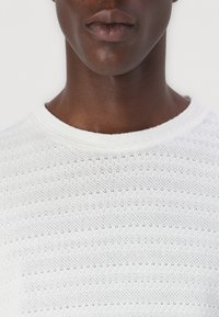 Hvit strikket genser med rund hals, med et teksturert mønster av små hull i horisontale striper. Glatt stoff med en lett følelse.