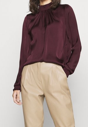 Kvinde iført en vinrød bluse med lange ærmer med opsamlede manchetter og beige højtaljede plisserede bukser, stående mod en ensfarvet baggrund.