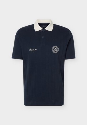 Πόλο μπλουζάκι σε σκούρο μπλε με κοντά μανίκια, λευκό γιακά, κουμπιά και το κείμενο «It’s on us. Since 1967» καθώς και κυκλικό λογότυπο στο στήθος.