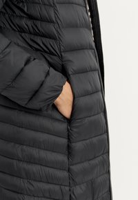 Sort pufferjakke med lodret søm, lynlåslukning og en sidelomme. Stoffet ser glat ud med en let glans.