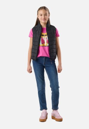 Dziewczyna stoi, ubrana w czarną pikowaną kamizelkę, różowy t-shirt z grafiką kota, niebieskie dżinsy i różowe trampki z gumowymi podeszwami.