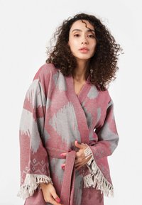 Pletený kúpeľový plášť v ružovej a šedej farbe s geometrickými vzormi, širokými rukávmi, opaskom na viazanie a strapcovými detailmi na manžetách. Mäkka textúra.