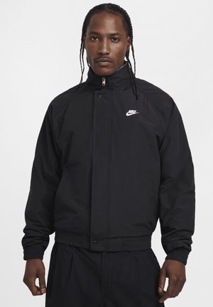 Zwarte nylon jas met hoge kraag, ritssluiting, elastische manchetten en een wit Nike-logo op de borst. Soepele textuur, relaxte pasvorm.