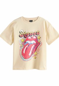 Vaaleankeltainen puuvillapaita, jossa on kuva punaisesta kielestä ja "Stones"-teksti rohkealla, tyylitellyllä fontilla, tähdillä korostettuna.