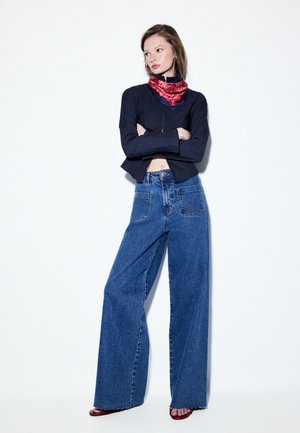 Młoda kobieta stoi ze skrzyżowanymi ramionami, ubrana w szerokie niebieskie dżinsy, czarną bluzkę z długim rękawem, czerwony wzorzysty szalik na szyi i czerwone sandały.