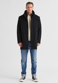 Schwarzer Wollmischungs-Mantel mit hohem Kragen, vorderer Knopfschluss und seitlichen Taschen, getragen über einem cremfarbenen Pullover und blauen Jeans.