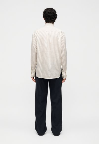 Άτομο με σγουρά μαλλιά που φοράει μακρυμάνικο, ανοιχτόχρωμο ριγέ πουκάμισο και φαρδιά μαύρη παντελόνα, στέκεται με την πλάτη σε απλό φόντο.