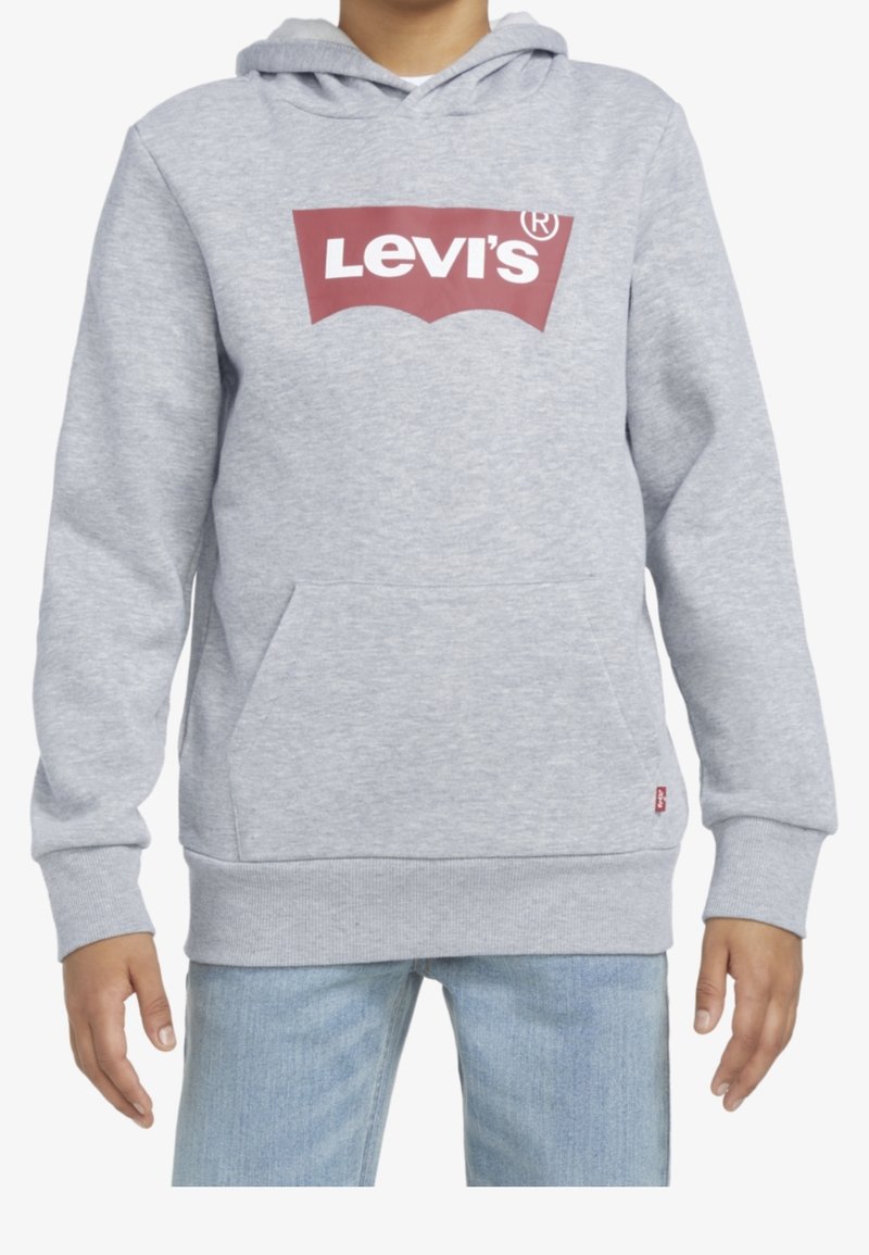 Enfant portant un sweat à capuche gris clair avec un logo Levi's rouge sur la poitrine et un jean bleu clair, mains détendues sur les côtés.
