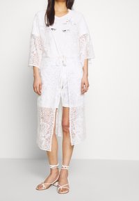 Kimono di pizzo bianco con motivo floreale, apertura frontale, maniche larghe e vita legata. Indossato sopra una t-shirt bianca e pantaloncini chiari. Sandali con cinturini.