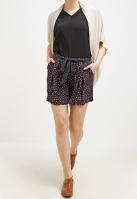 Czarna bluzka z dekoltem w serek połączona z granatowymi szortami w czerwono-białą kropkowaną wzór, przewiązany w talii pasującym paskiem. Brązowe sznurowane buty.