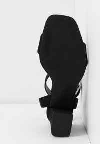 Sandale pour femme en daim noir avec talon bloc, bout carré, bride à la cheville et boucle argentée, vue de la semelle sur fond blanc.