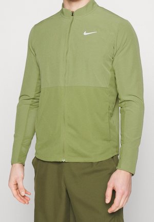 Man wearing light green zip-up Nike jacket and dark green pants, standing with arms relaxed by sides.