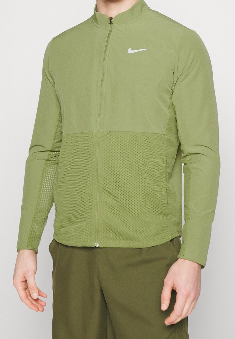 Homme portant une veste Nike zippée vert clair et un pantalon vert foncé, debout avec les bras détendus le long du corps.