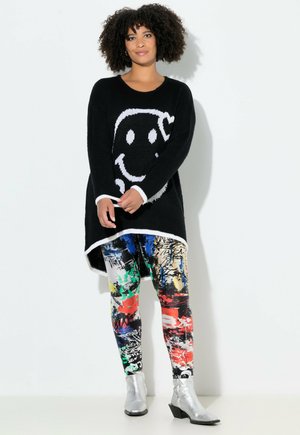 Maglione nero lavorato a maglia con grafica di un volto sorridente bianco, abbinato a leggings con un motivo astratto colorato. Stivali d'argento alla caviglia completano l'outfit.