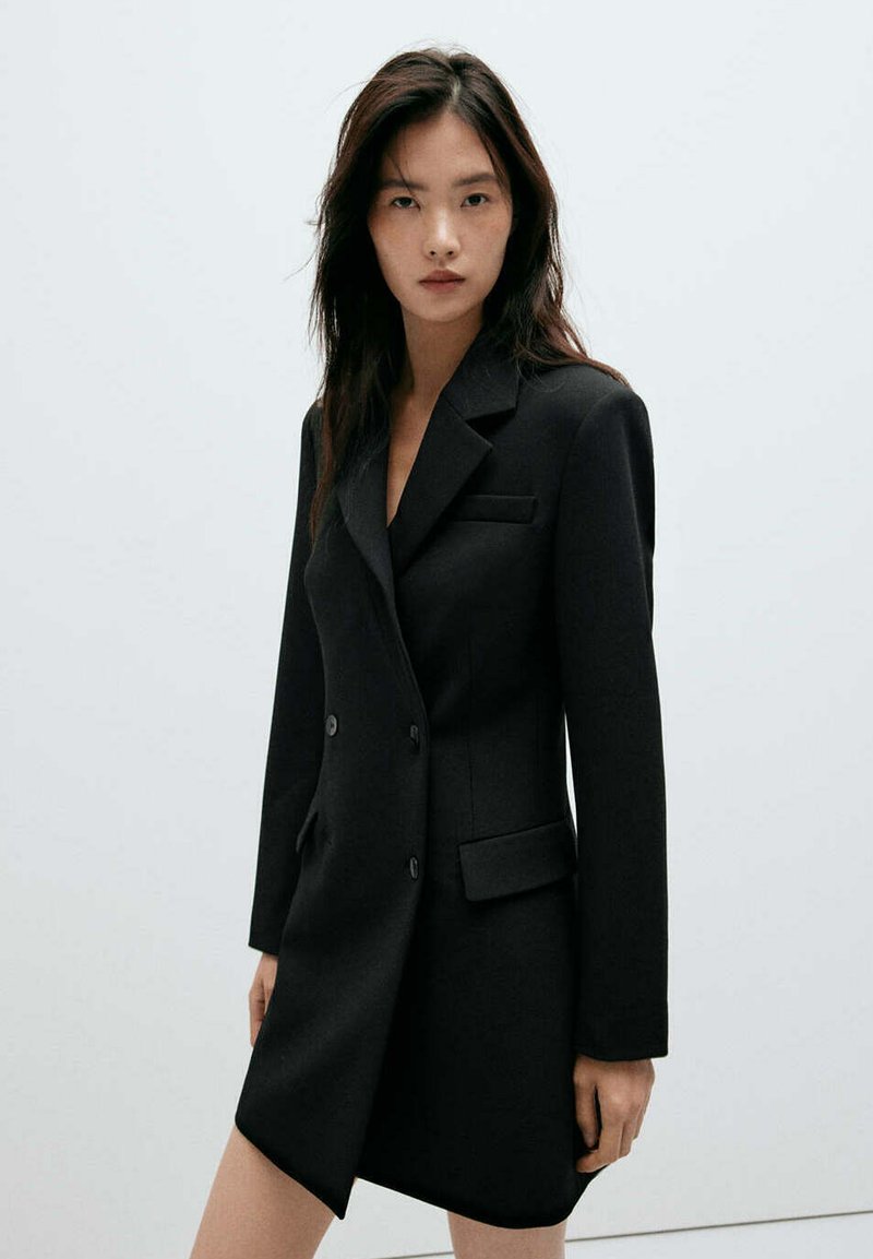 Jeune femme debout devant un fond uni, portant une robe blazer noire ajustée à double boutonnage avec des manches longues.