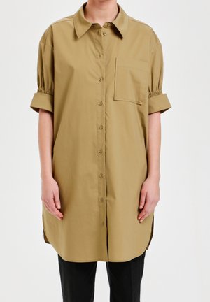 Osoba nosząca luźną, sięgającą kolan, beżową sukienkę koszulową na guziki z podwiniętymi rękawami i kieszonką na piersi, w zestawieniu z czarnymi spodniami.