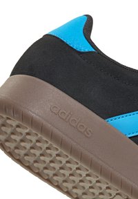 Nahaufnahme einer schwarzen und blauen Sneaker-Ferse mit strukturierter brauner Sohle und eingeprägtem Adidas-Logo.