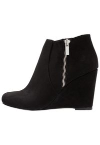 Czarny zamszowy botek na koturnie z zamkiem błyskawicznym z boku i zaokrąglonym noskiem, pokazany z zewnętrznej strony na białym tle.