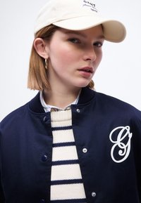 Jeune femme aux cheveux courts portant une casquette beige, une boucle d'oreille en or en forme d'anneau, une veste bleu marine avec un monogramme blanc, et un pull en tricot rayé sur fond blanc.