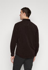 Camisa de pana marrón oscuro con mangas largas, puños abotonados y cuello extendido. Presenta un dobladillo curvado y una tela texturizada.