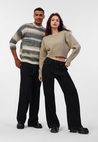 Deux mannequins portent des tenues décontractées : un pull en maille rayé vert, beige et gris ; un pull beige raccourci ; un jean noir à jambes larges ; des bottes noires imposantes.