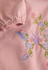 Un tissu rose avec une finition texturée présente des broderies colorées de fleurs et de papillons. Les détails des poignets élastiqués mettent en valeur le design.