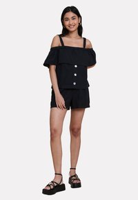 Top nero scoperto sulle spalle con strato arricciato, chiusura frontale con bottoni e pantaloni corti neri abbinati. Il modello indossa sandali con plateau neri.