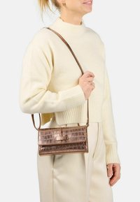 Donna che indossa un maglione crema e pantaloni, mentre porta una piccola borsa a tracolla marrone con texture al coccodrillo.