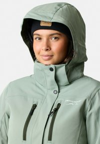 Jasnozielona kurtka typu softshell z kapturem, zapinanymi kieszeniami i logo na klatce piersiowej. Charakteryzuje się gładką fakturą i dopasowanym krojem.