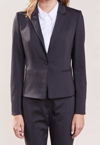 Veste noire ajustée avec une fermeture à un bouton, des revers crantés et deux poches avant, associée à une chemise blanche à col en dessous.