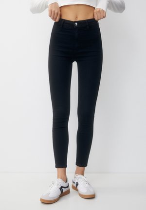 Personne portant un jean skinny noir taille haute et des baskets blanches avec des rayures noires, debout devant un fond blanc uni.
