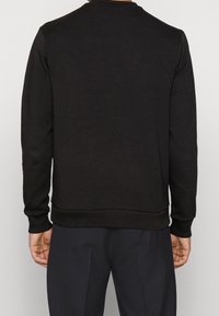 En person som bär en svart sweatshirt, sedd bakifrån, med en enfärgad bakgrund. Deras händer syns vid sidorna.