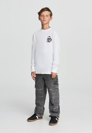 Camicia bianca a manica lunga con un design di serpente nero sul petto, abbinata a pantaloni cargo grigi e scarpe da ginnastica nere con dettagli bianchi.