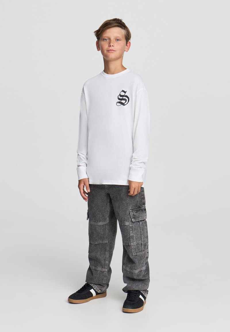 Biała koszula z długim rękawem z czarnym wzorem węża na klatce piersiowej, w połączeniu z szarymi spodniami cargo i czarnymi sneakersami z białymi akcentami.