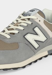 Baskets New Balance 574 Classic grises et beiges avec lacets blancs, logo "N" blanc et semelle texturée visible sur un fond clair.