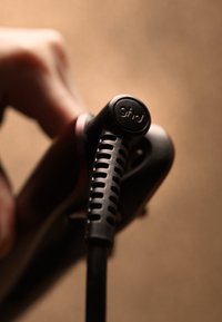 Grepp till svart hårplattång med texturerad, räfflad sladd. Logotyp synlig på en slät yta, subtila metalliska accenter. Brun bakgrund.