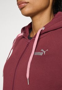 Burgundia värvi tõmblukuga kapuutsiga fliis, millel on roosad paelad ja hall Puma logo. Pehme kangas sileda tekstuuriga ning kapuuts.