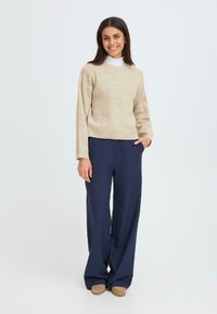 Bežový rebrovaný sveter s dlhými rukávmi, doplnený námorníckymi širokými nohavicami. Doplňky zahŕňajú bielu košeľu s golierom, ktorá je navrstvená pod sveter.