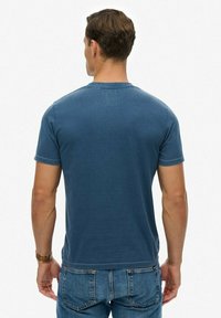 Blauw katoenen T-shirt met een ronde hals, korte mouwen en zichtbare stiksel details. Draagt blauw spijkerbroek. Achteraanzicht, met nadruk op de pasvorm van het shirt.