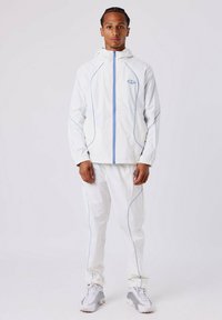 Jeune homme se tenant debout, portant un survêtement à capuche blanc avec des bordures bleues, associé à des baskets blanches et grises sur un fond uni.