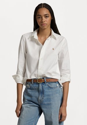 White cotton long-sleeve shirt with a button-down collar and a small embroidered logo on the chest, paired with light blue distressed jeans and a brown belt.