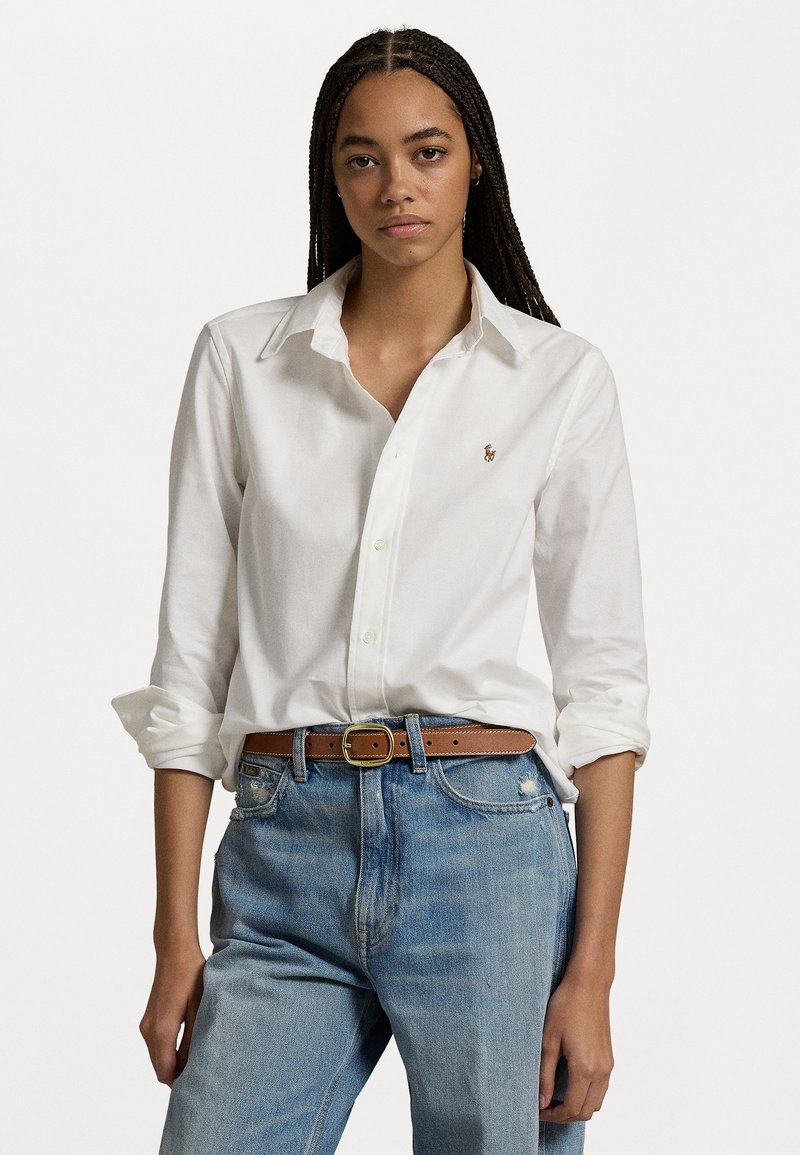 Hvit langermet skjorte i bomull med krage med knapper og en liten brodert logo på brystet, kombinert med lyseblå slitte jeans og et brunt belte.